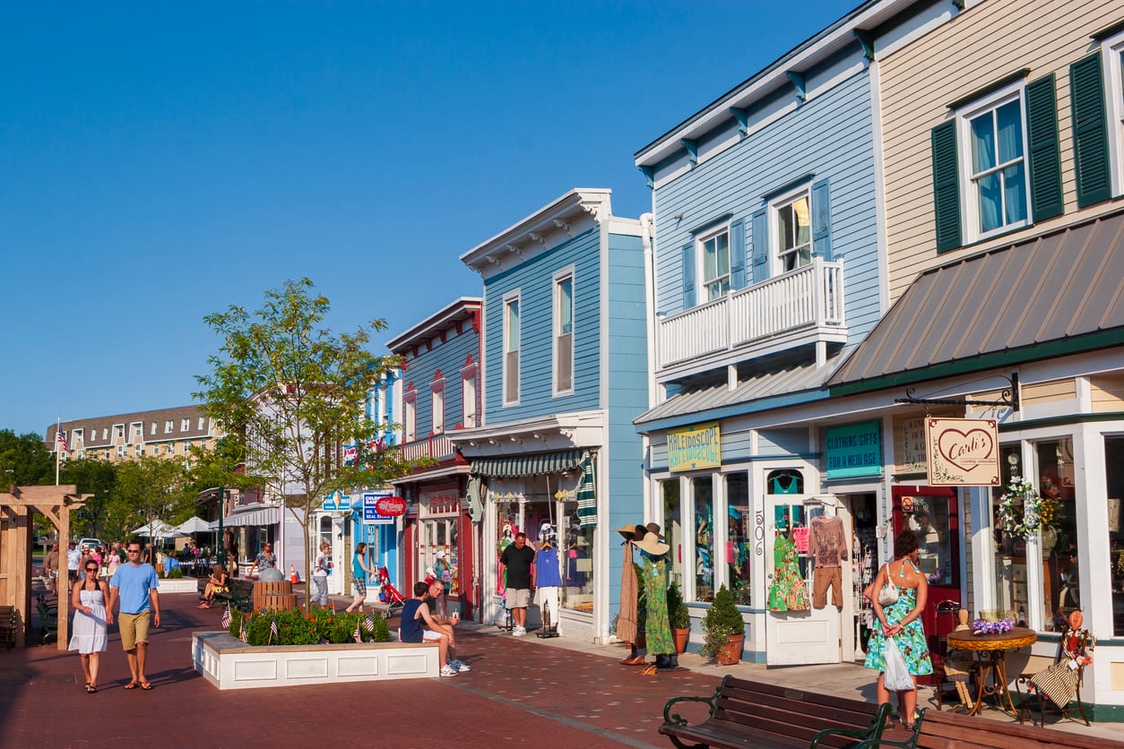 People stroll and shop along a sunny, colorful street lined with boutique stores, benches, and flower planters on a clear day.
