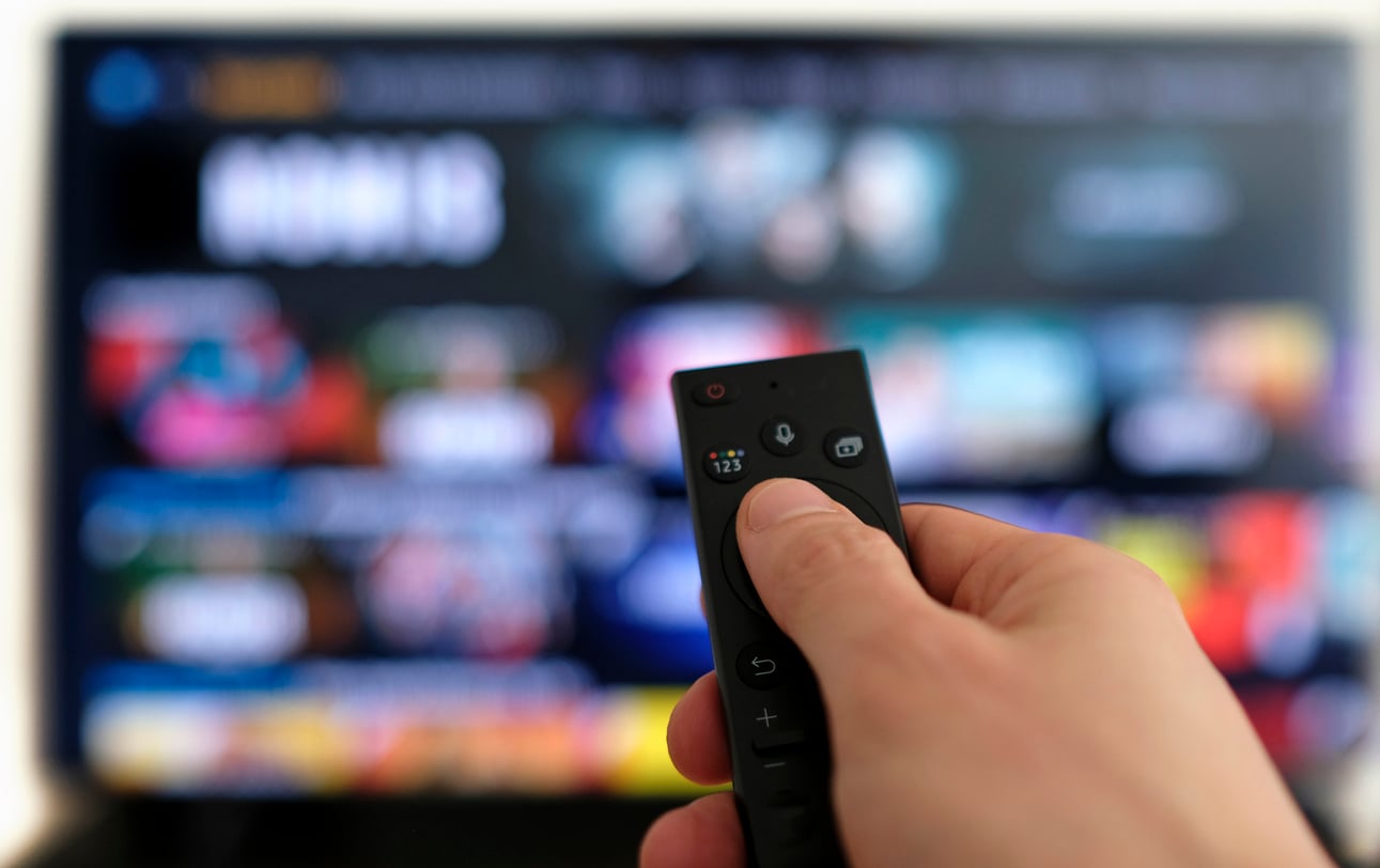 A hand holds a black remote control pointed toward a blurred television screen displaying colorful streaming service options and thumbnails, highlighting how some viewers feel overwhelmed by waste of money products scattered across countless platforms.