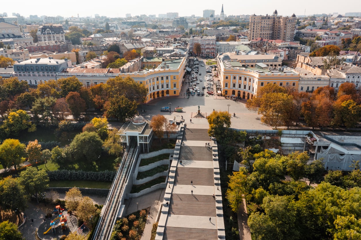 Aerial view of a wide staircase surrounded by green trees leading to a city square with curved yellow buildings, busy streets, and a mix of historic and modern architecture—one of the city's must-see travel destinations.