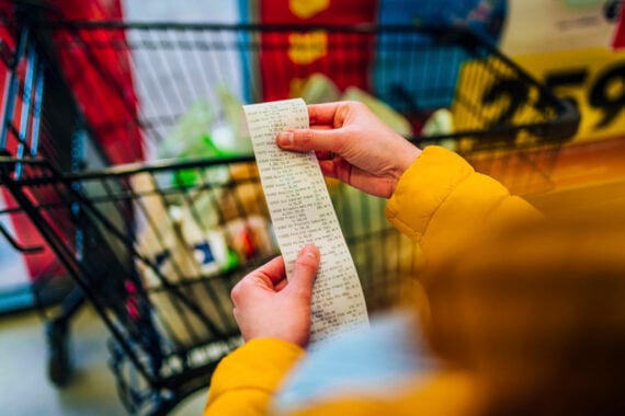 A person wearing a yellow jacket holds a long receipt in front of a shopping cart filled with groceries at a store.