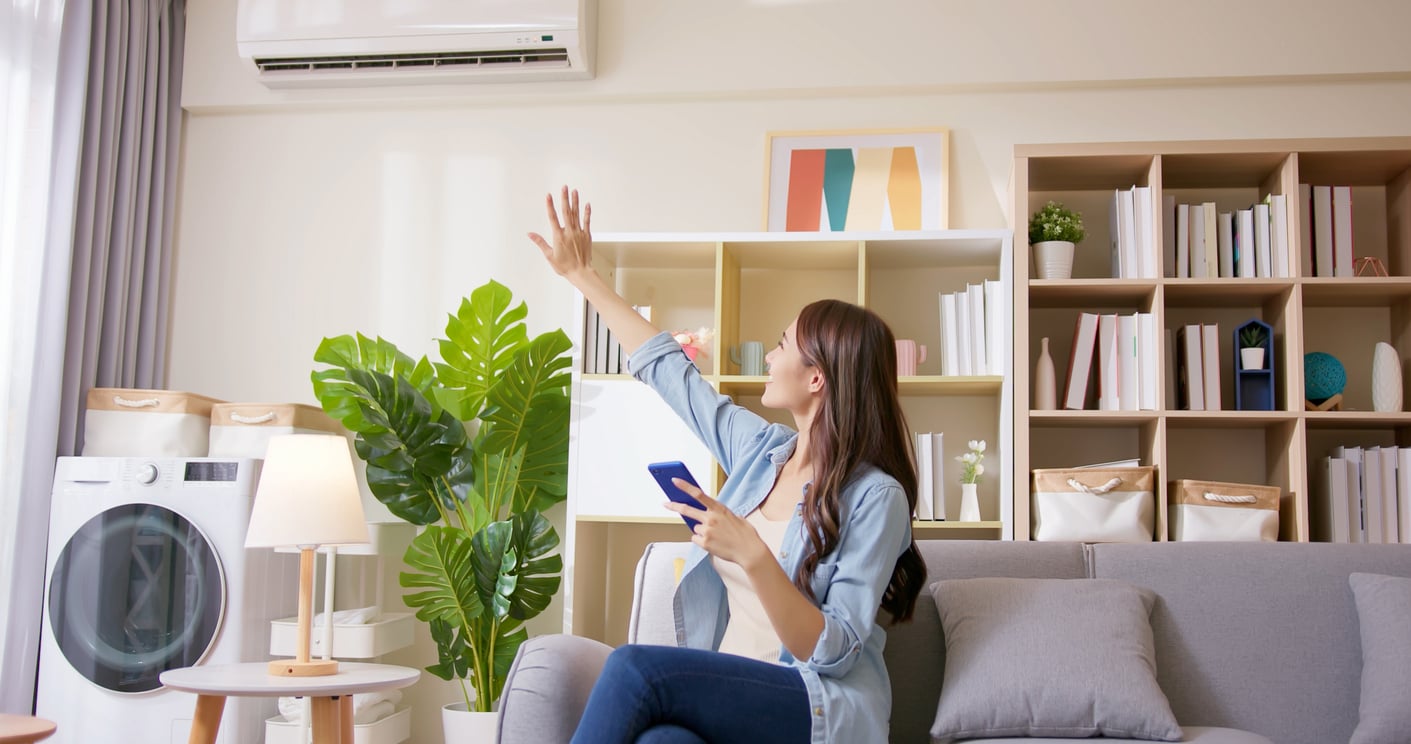 A woman sits on a couch holding a phone and raises her hand toward an air conditioner on the wall in a bright living room with modern decor, bookshelves, plants, and a washing machine, thoughtfully managing her comfort while keeping an eye on monthly bills.