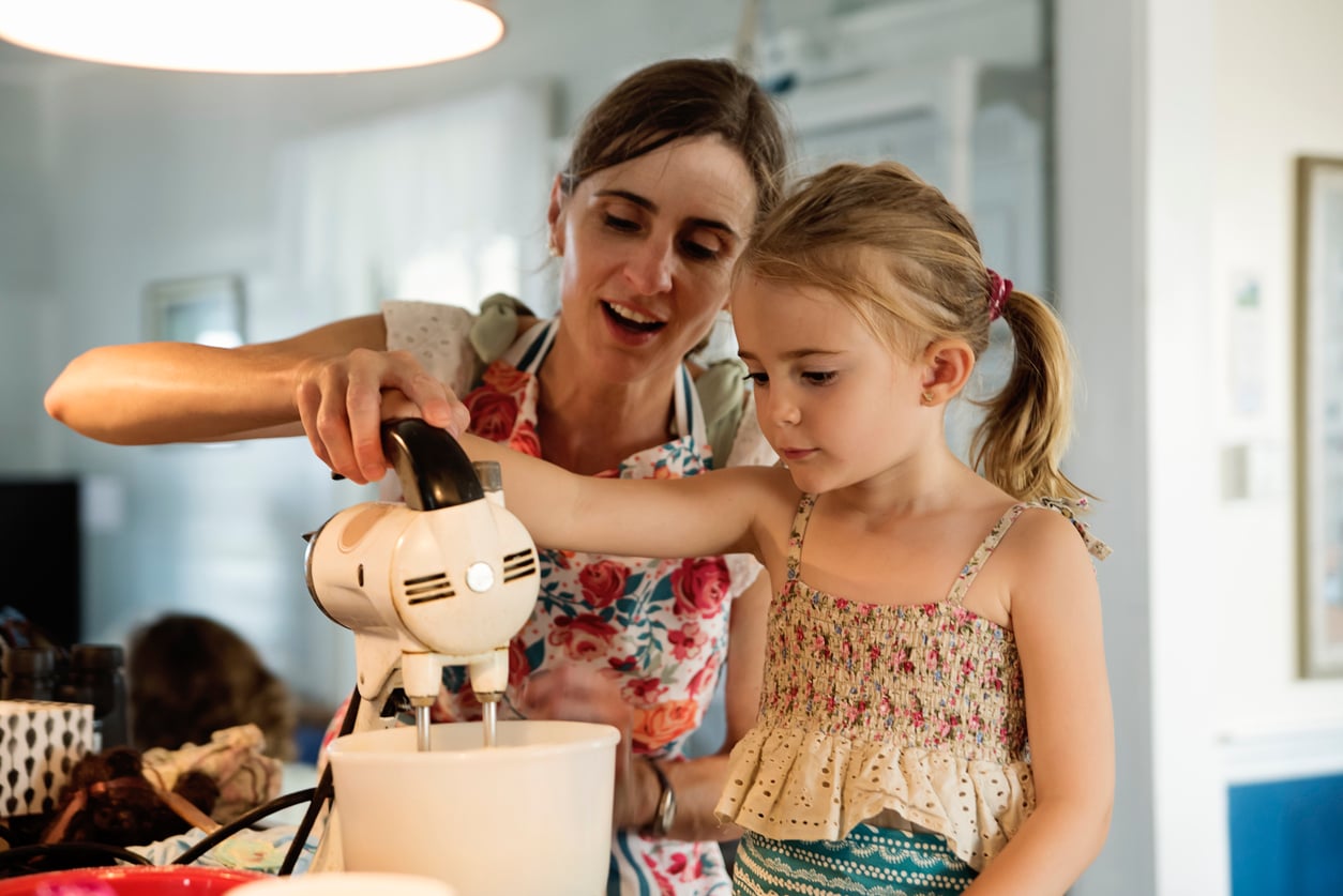 A woman and a young girl use a hand mixer together in a bright kitchen, mixing ingredients in a white bowl while smiling and wearing casual clothes, surrounded by various kitchen items.