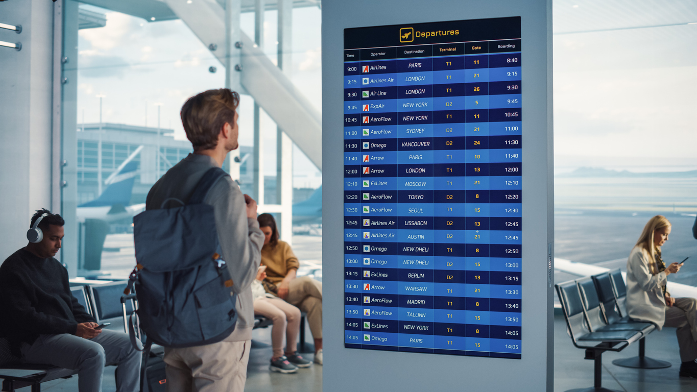 A man with a backpack stands in an airport terminal, looking at a large electronic departures board. Other travelers sit nearby, some using their phones. Large windows show airplanes and the runway outside.
