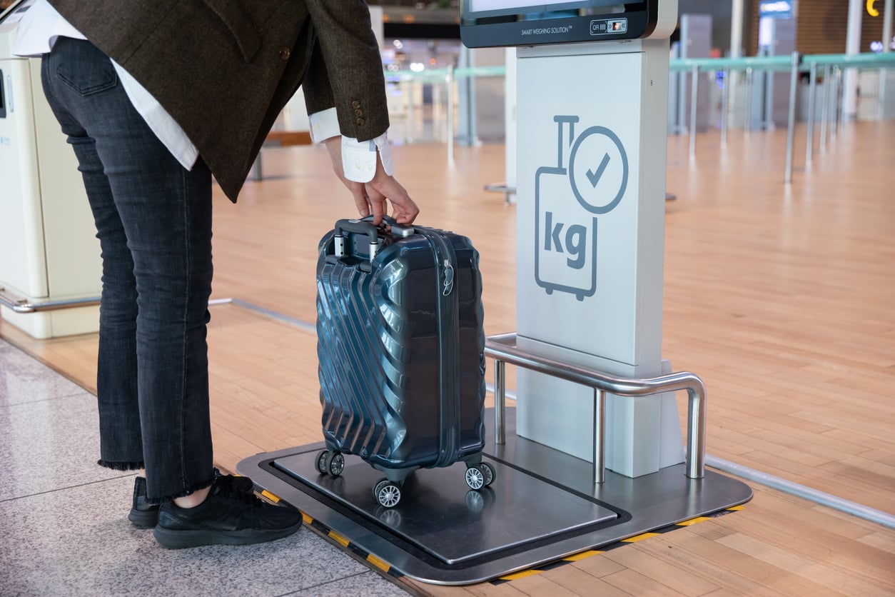 A person places a small suitcase on a luggage scale at an airport check-in area, ensuring the bag meets weight requirements before booking a flight. The scale has a sign with a suitcase and "kg" written on it.
