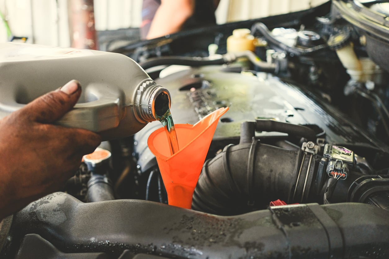 A person pours engine oil from a gray bottle into an orange funnel placed in a car engine bay, performing routine vehicle maintenance as part of their road trip checklist.