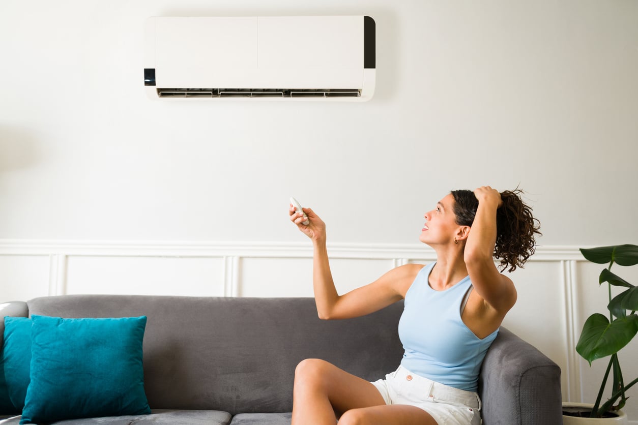A woman sitting on a gray sofa points a remote control at an air conditioner on the wall, looking up while holding her hair, with blue cushions and a plant beside her.