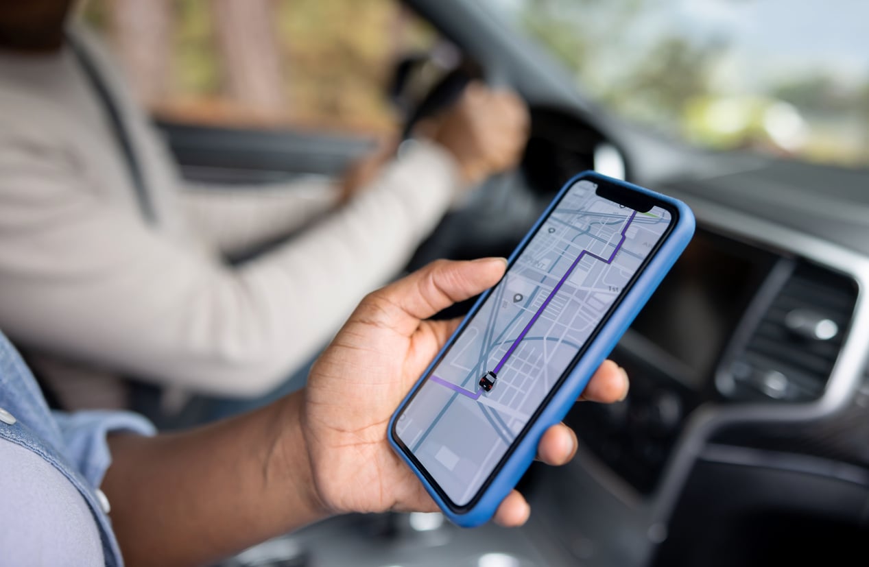 A person in a car holds a smartphone displaying a navigation app with a route on the map, while another person drives.