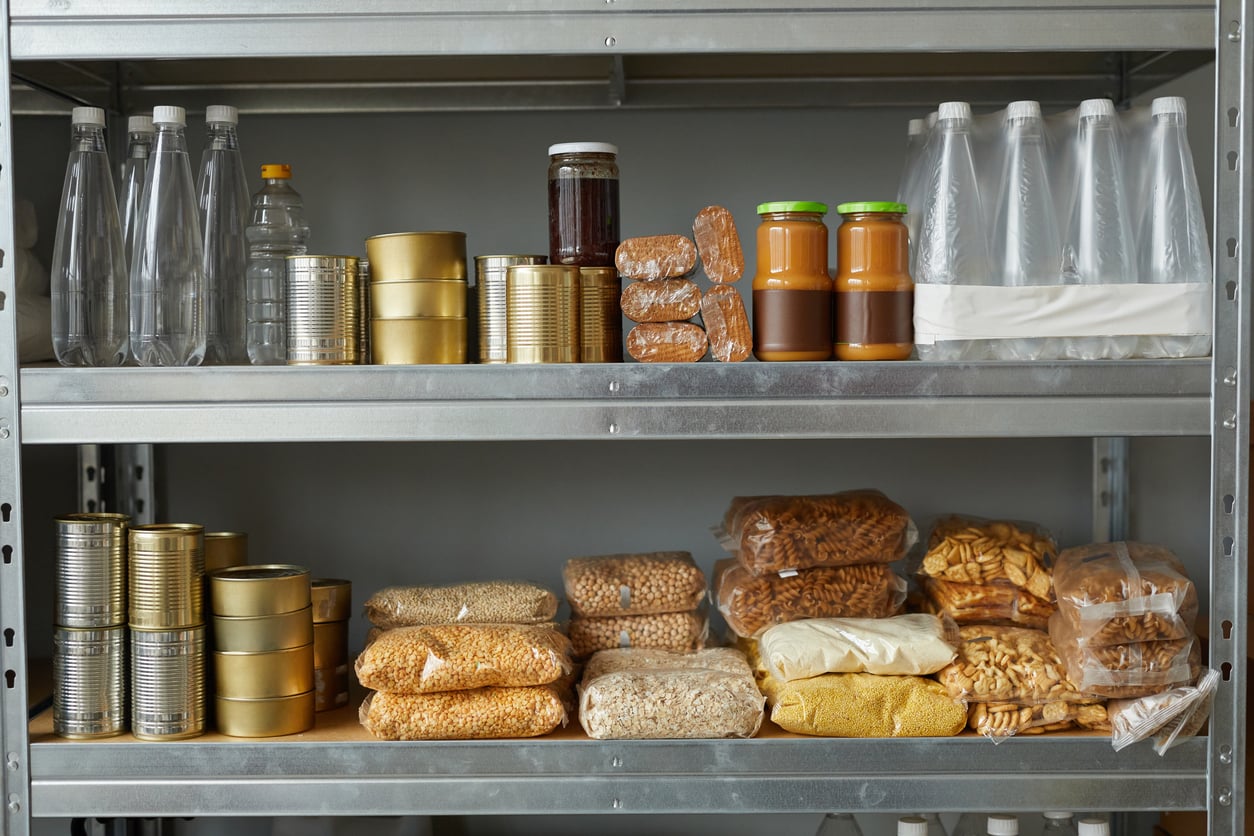 Metal shelves, reminiscent of a well-organized Walmart aisle, are stocked with canned food, bottled drinks, jars, and plastic bags containing grains, pasta, and dried food items—all neatly arranged in a pantry setting.
