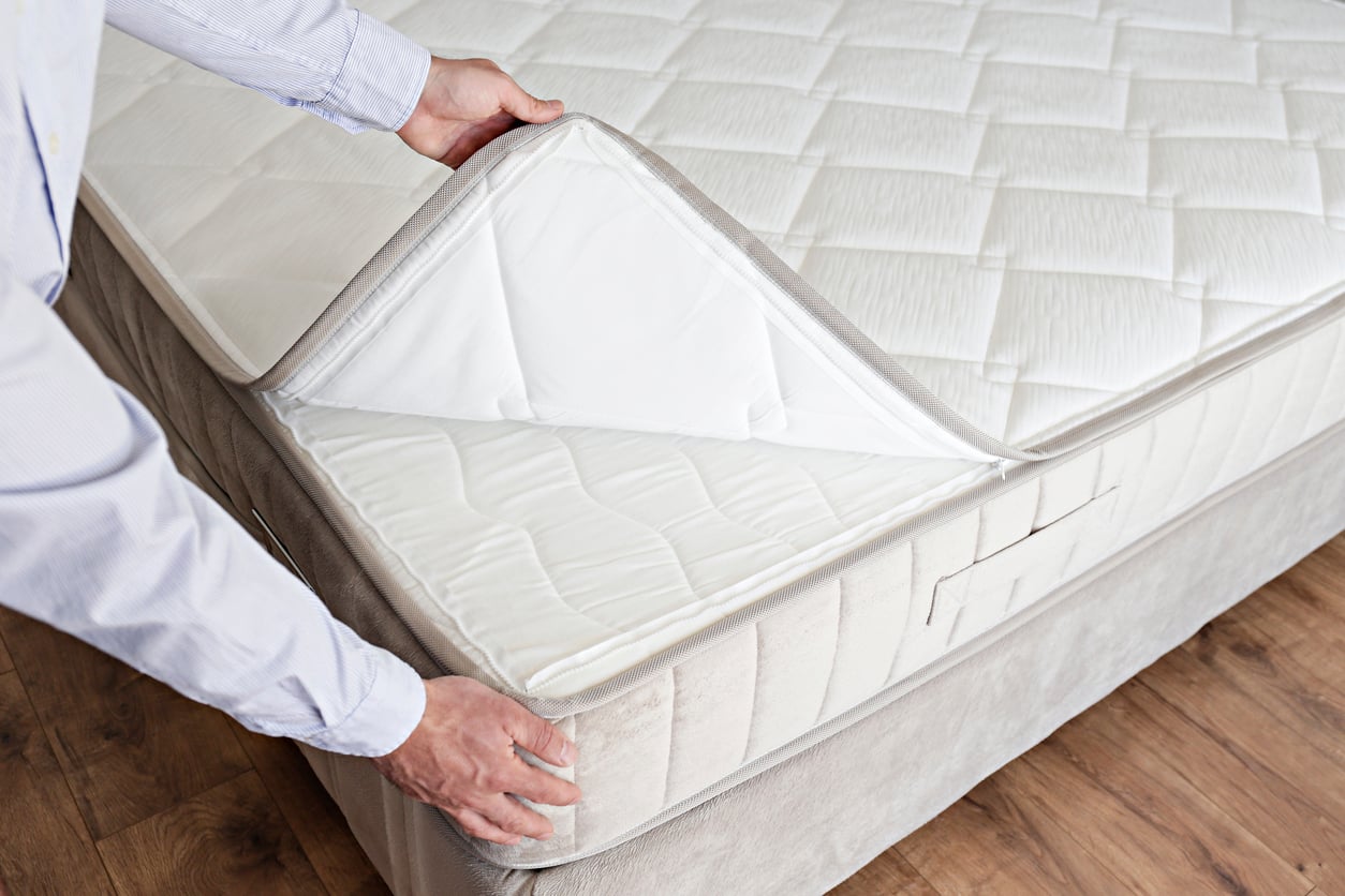 A person lifts the corner of a mattress protector to reveal the products beneath. The bed has a quilted white cover and sits on a wooden floor.