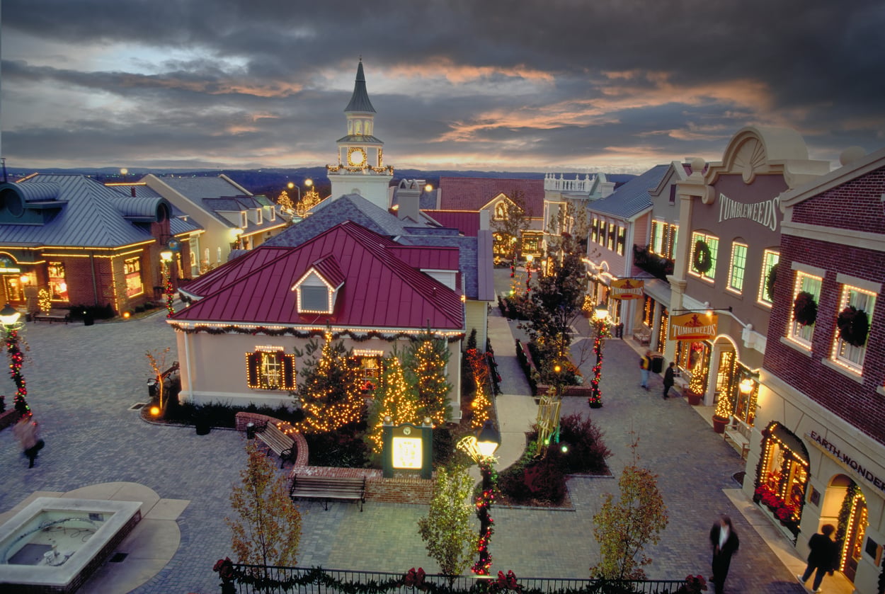 A festive shopping village at sunset, decorated with holiday lights and garlands, featuring charming buildings, a clock tower, and a dramatic cloudy sky overhead—just like one of the best Christmas towns in America.