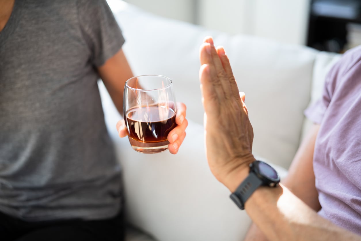 Two people sit on a couch. One person offers a glass of dark liquid while the other raises a hand in refusal, indicating they do not want the drink—perhaps demonstrating their money saving habits by choosing not to indulge.