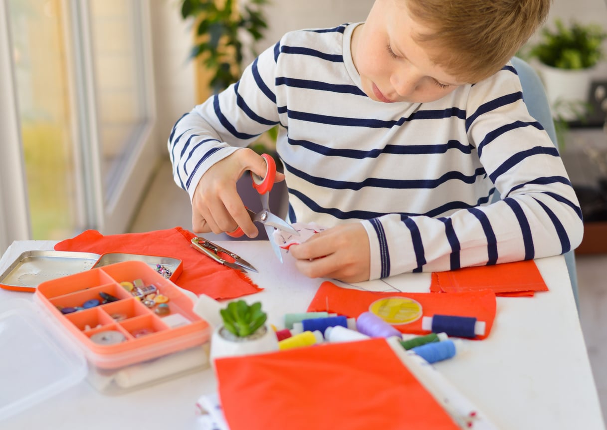 A young boy in a striped shirt uses scissors to cut white fabric at a table with sewing supplies, thread spools, buttons, and orange cloth pieces.