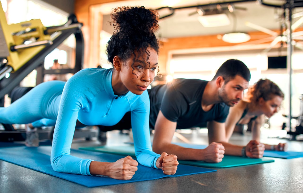 Three people exercise side by side on yoga mats in a gym, holding plank positions. The focus is on a woman in a blue outfit, with a man and another woman behind her—proving you can save money by working out together using shared gym equipment.