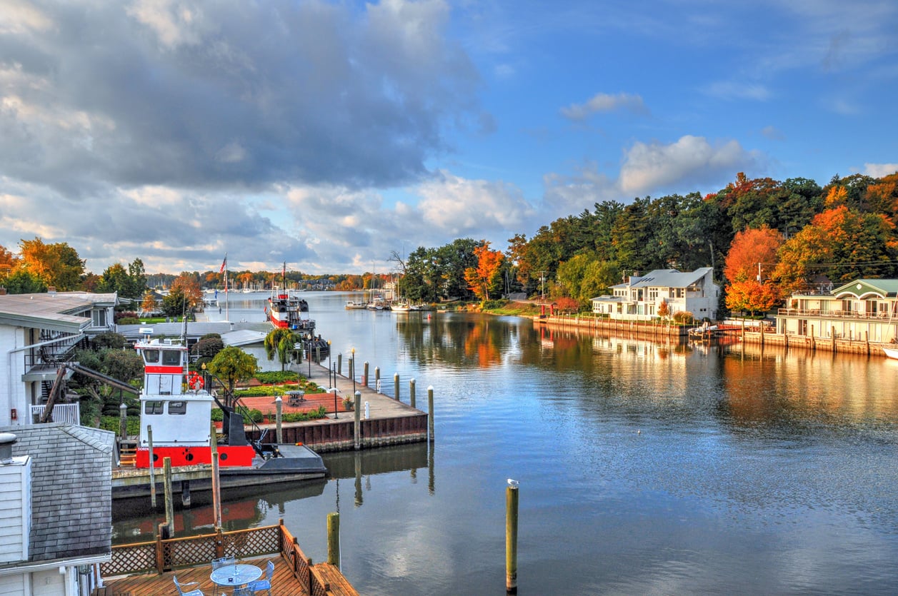 A calm river lined with docks, boats, and houses on both sides, surrounded by trees with autumn foliage under a partly cloudy blue sky.