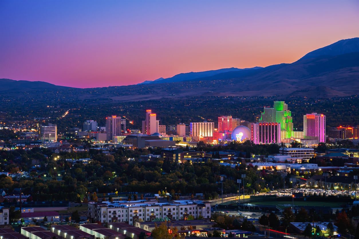 Cityscape at dusk with colorful neon lights on buildings, mountains in the background, and a gradient sky transitioning from orange to purple.