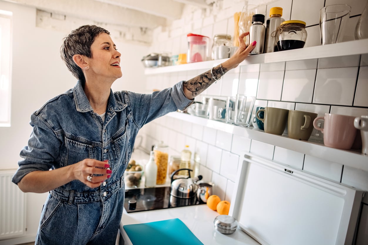 A smiling person in a denim jumpsuit reaches for a jar on a kitchen shelf, surrounded by mugs, jars, and fruit, in a bright, modern kitchen free of the home features homeowners no longer want.