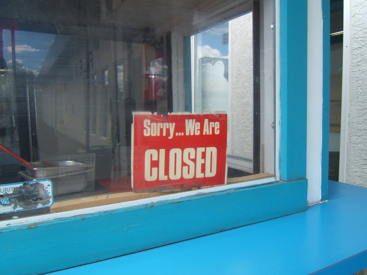 A red and white sign in a window reads “Sorry... We Are CLOSED.” The sign is behind glass, reflecting the sky and surrounding buildings. The window frame and counter are painted bright blue—a striking detail you might jot down while updating your road trip checklist.