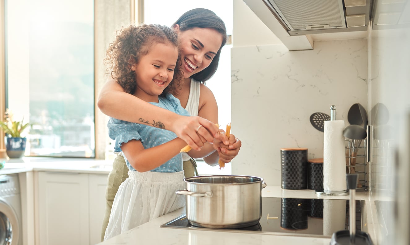 A smiling woman helps a young girl crack eggs into a pot on a stove in a bright, modern kitchen. Both appear happy and engaged in cooking together.
