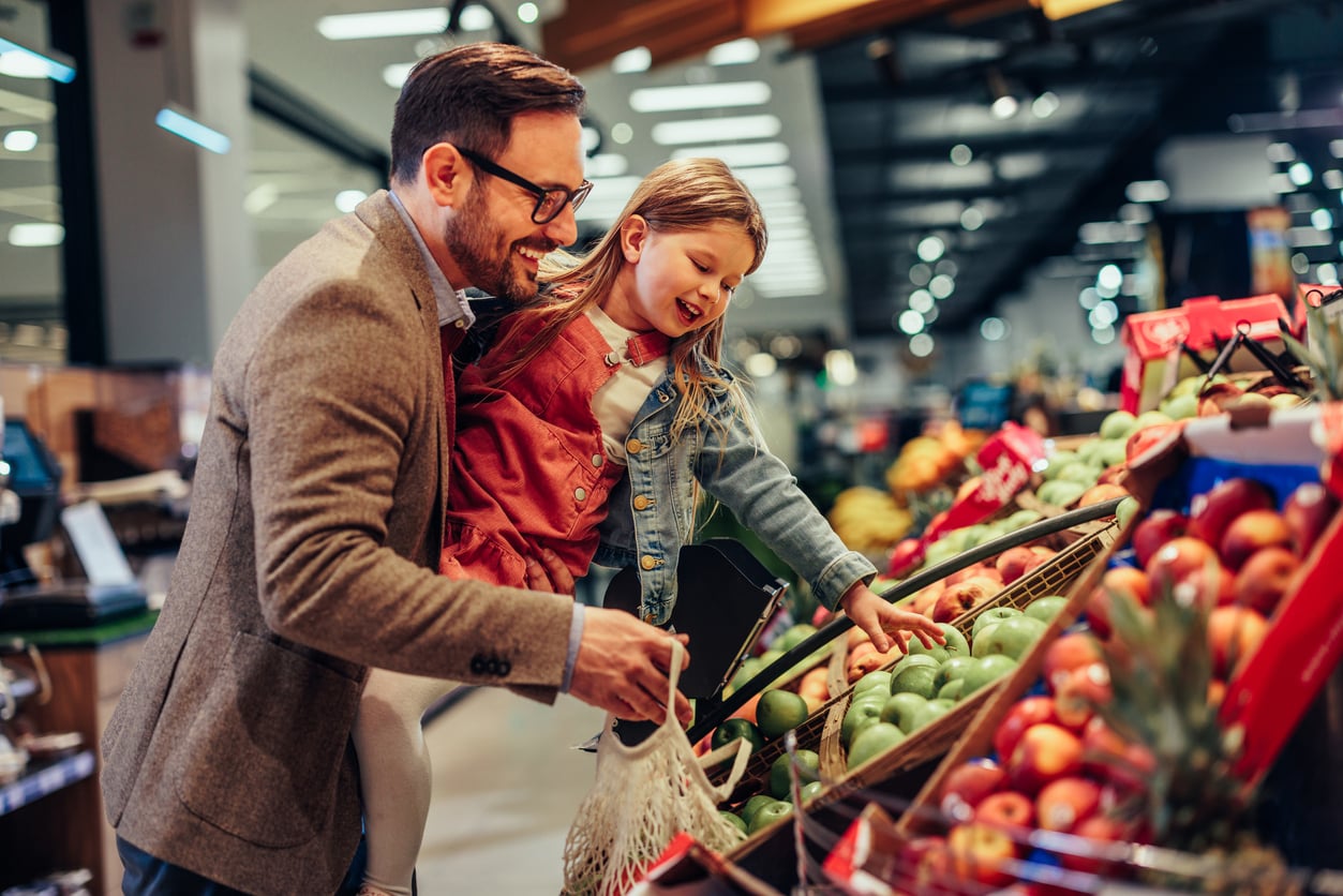 A man and a young girl smile while picking apples together in a grocery store's produce section, perhaps gathering fresh fruit for their vacation swaps adventure. The man holds a reusable bag as the girl reaches for fruit.