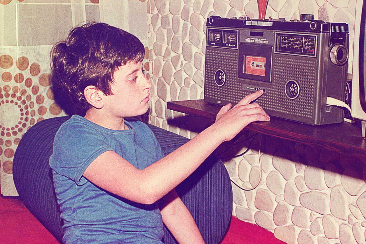 A boy in a blue shirt sits on a couch, pressing play on a vintage cassette boombox. The background features retro-patterned wallpaper and a curtain with circular designs.