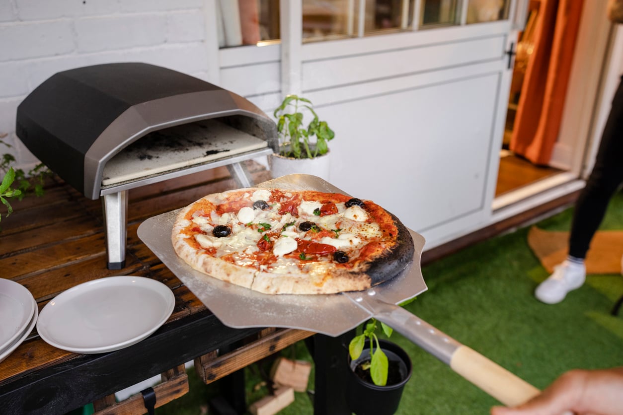 A freshly baked pizza with olives, cheese, and basil is being taken out of a small outdoor pizza oven using a pizza peel—an inviting scene that showcases the joy of outdoor home products. Plates and green plants are nearby.