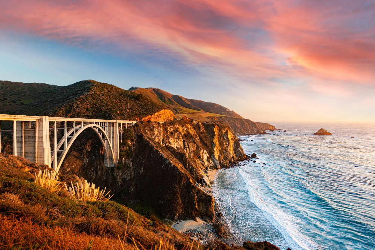 Bixby Creek Bridge spans rugged cliffs along California’s Big Sur coast at sunset, with waves crashing below and a colorful sky above.