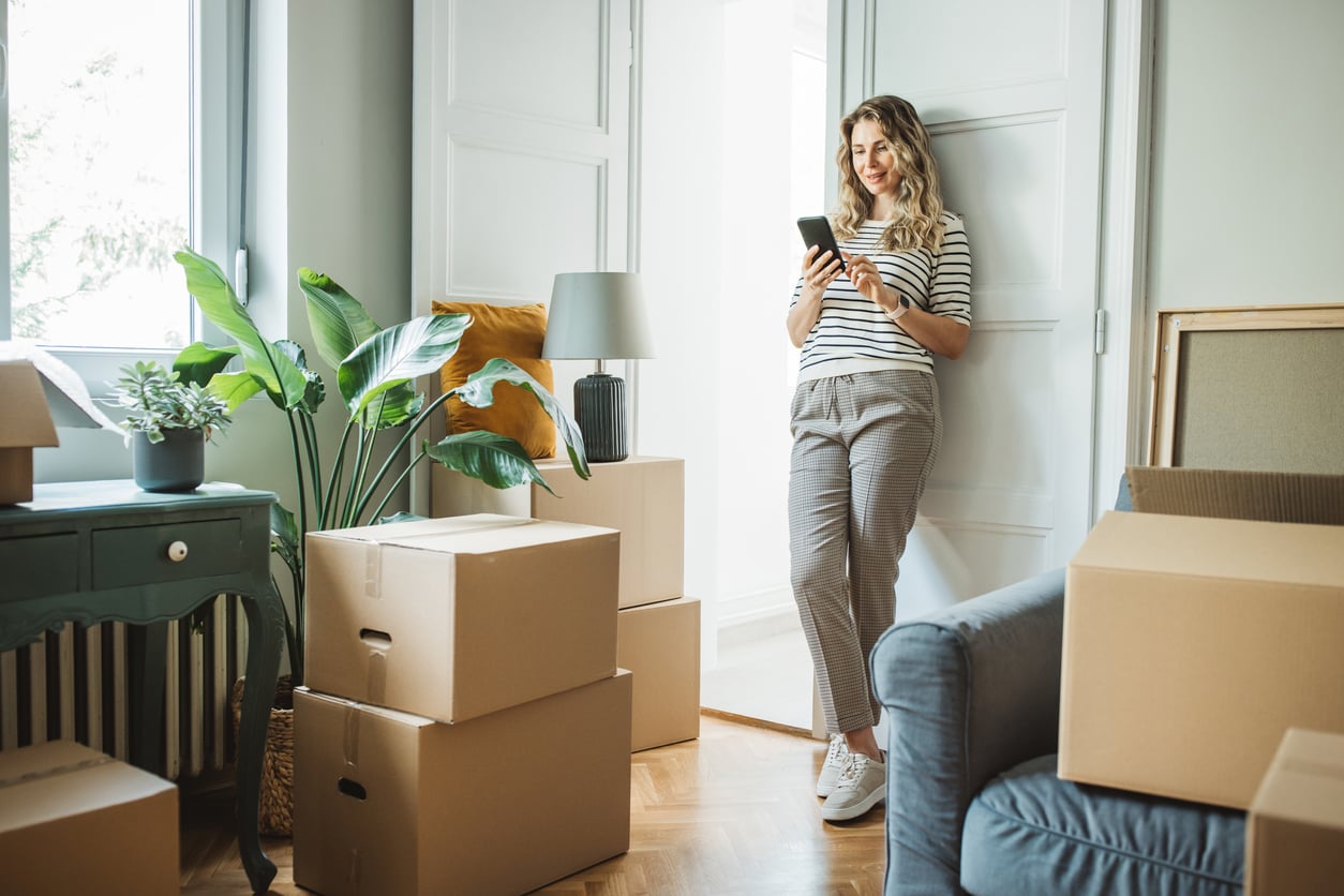 A woman in casual clothes leans against a door, smiling at her phone. She is surrounded by cardboard moving boxes, houseplants, and furniture in a bright, sunlit room.