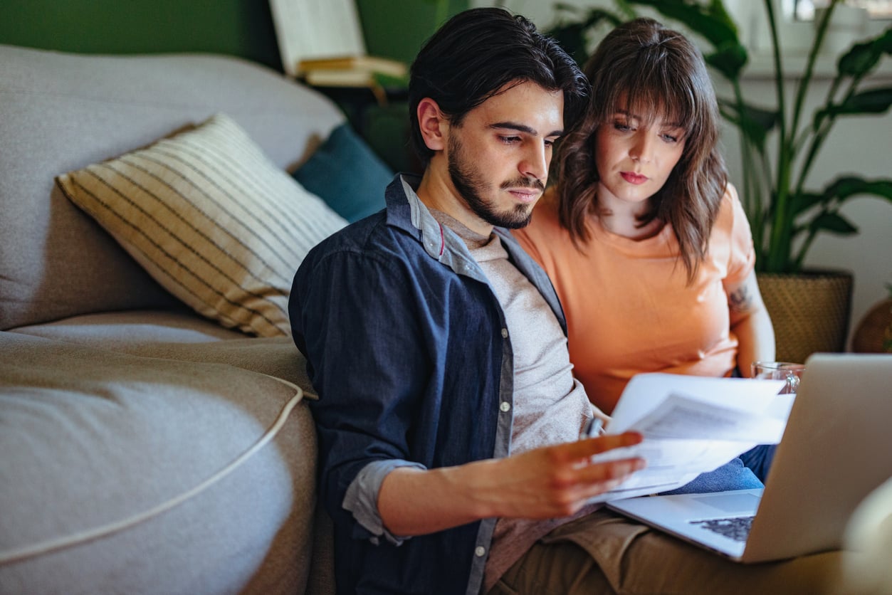 A couple sits on the floor by a couch, looking at documents together with a laptop in front of them. They appear focused and concerned, possibly reviewing bills or finances in a cozy living room.