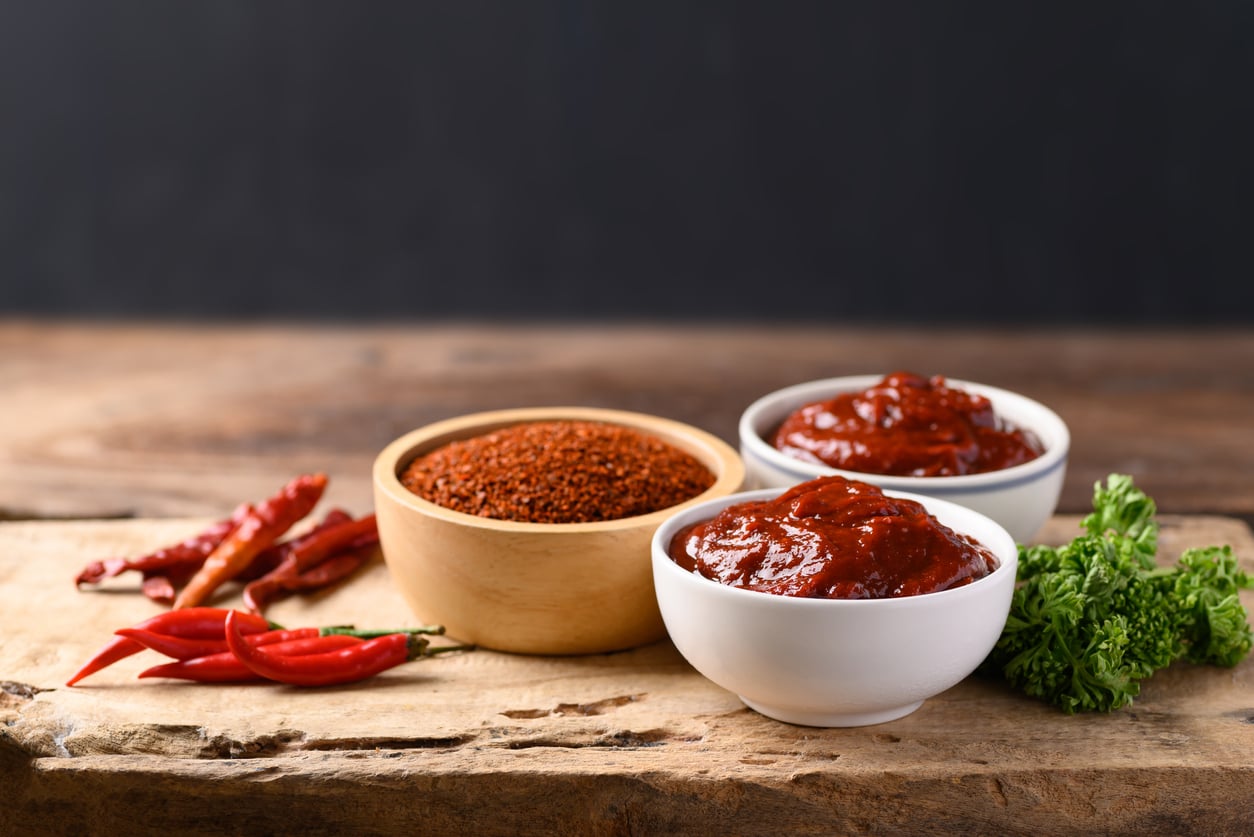 Two white bowls filled with red chili paste and a wooden bowl of red chili powder sit on a rustic wooden surface, with fresh red chilies and green parsley beside them.