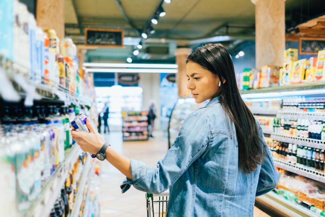 A woman in a denim jacket stands in a grocery store aisle, examining a product from the refrigerated section while holding a shopping cart.