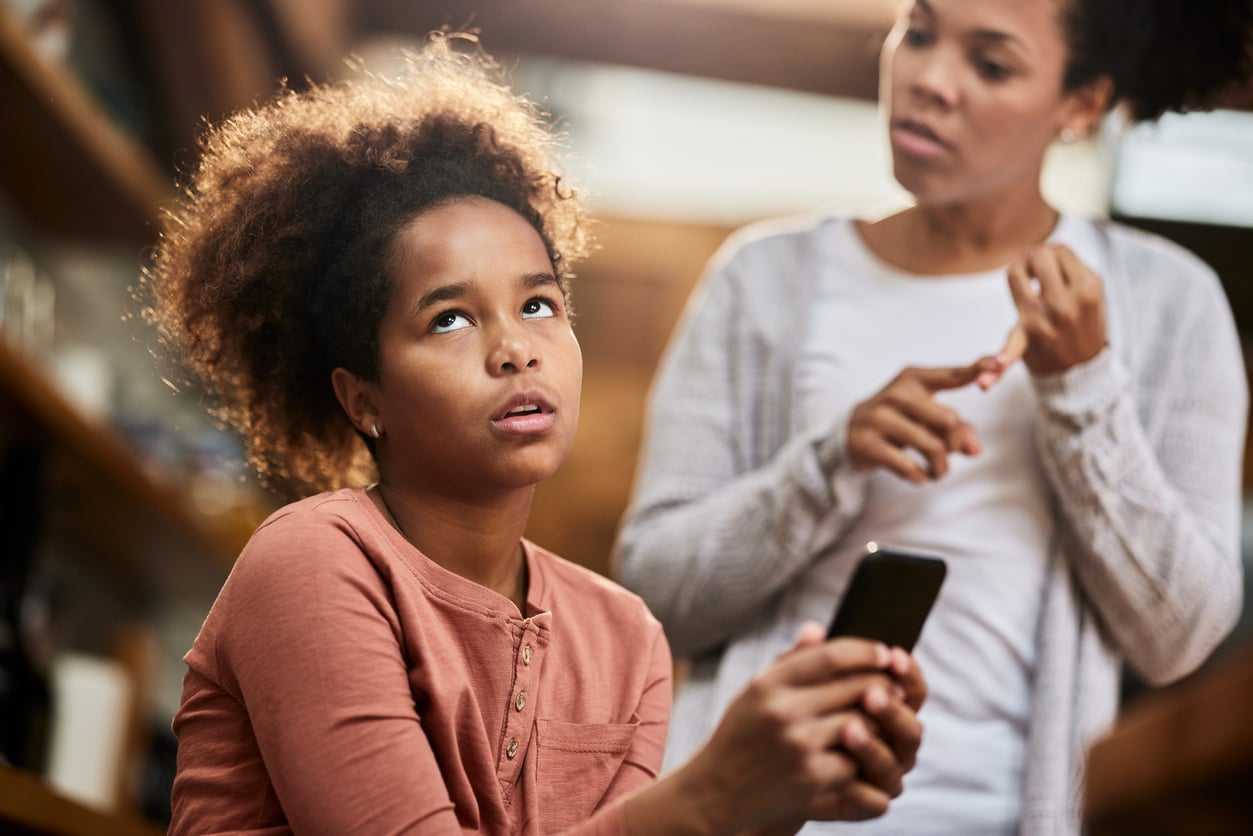 A young girl holding a smartphone rolls her eyes in frustration while an adult woman in the background appears to be talking to her.