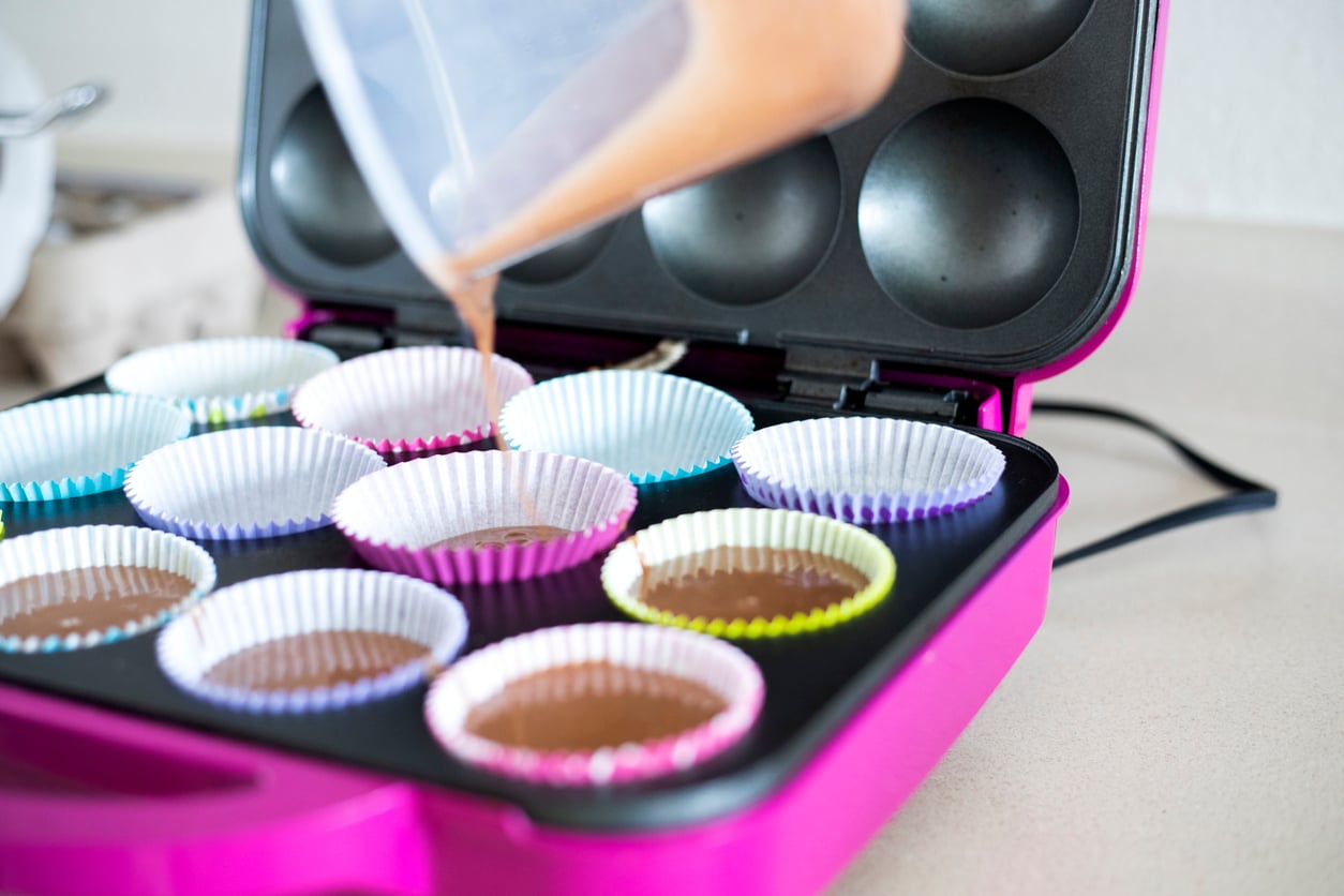 A close-up of cupcake batter being poured into colorful paper liners in a pink cupcake maker on a kitchen counter, showcasing one of the must-have kitchen gadgets for baking enthusiasts.