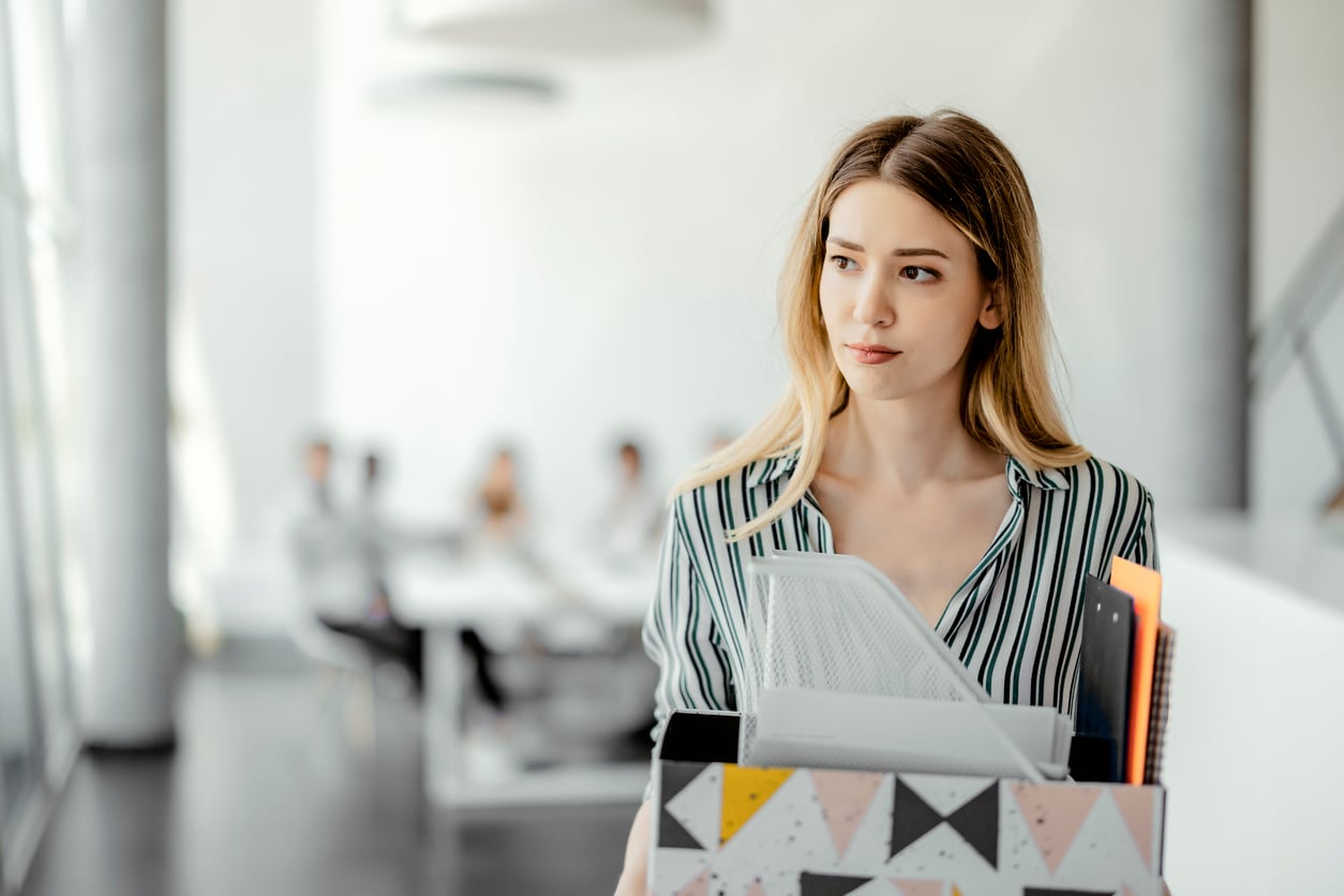 A woman with long blonde hair carries an office box filled with folders and supplies, looking thoughtful, while people sit at a conference table in the background of a bright office.