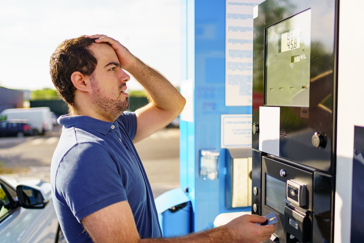 A man at a gas pump looks surprised and worried, holding his head with one hand while paying with the other. The fuel price is visible on the pump display, making him feel like fueling up has turned into yet another waste of money product.