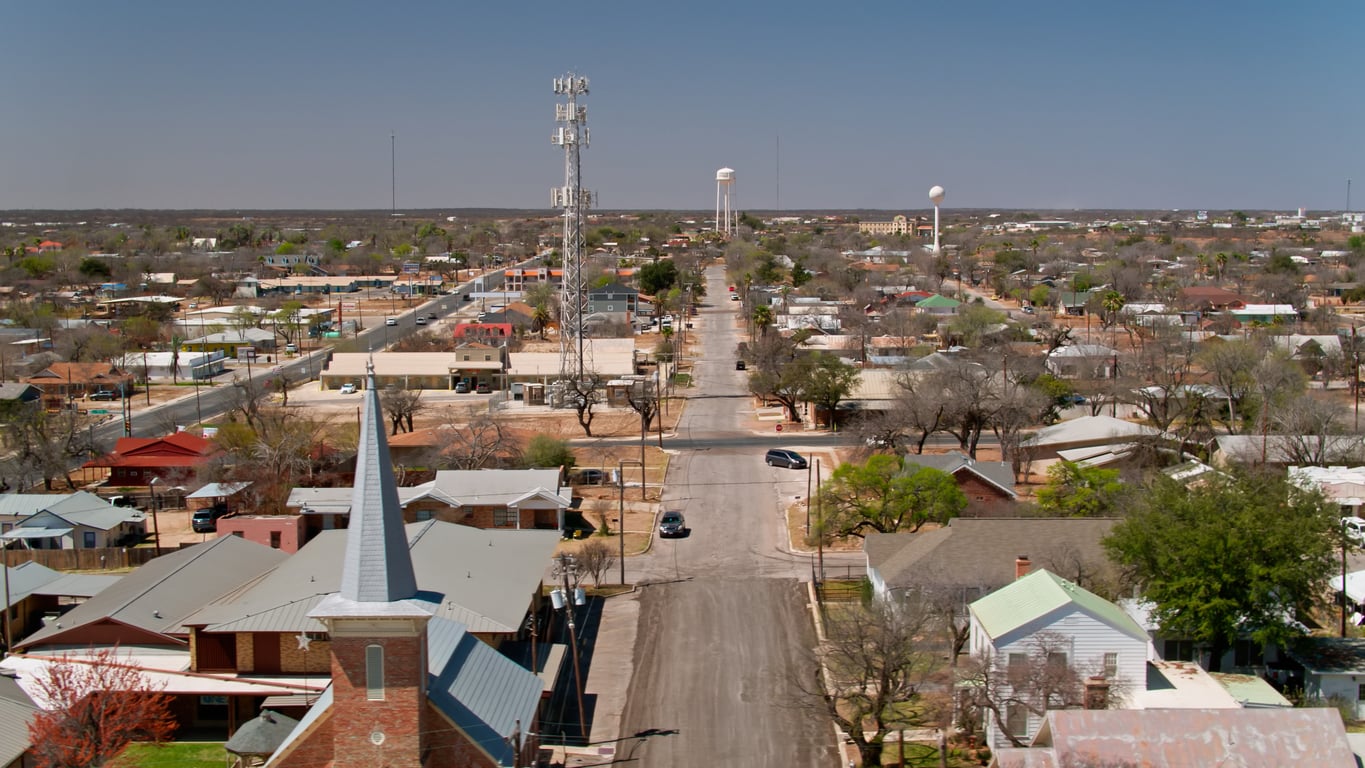 A small town reminiscent of classic American towns, with a church in the foreground, tree-lined streets, houses, water towers, and a tall cell tower under a clear blue sky.