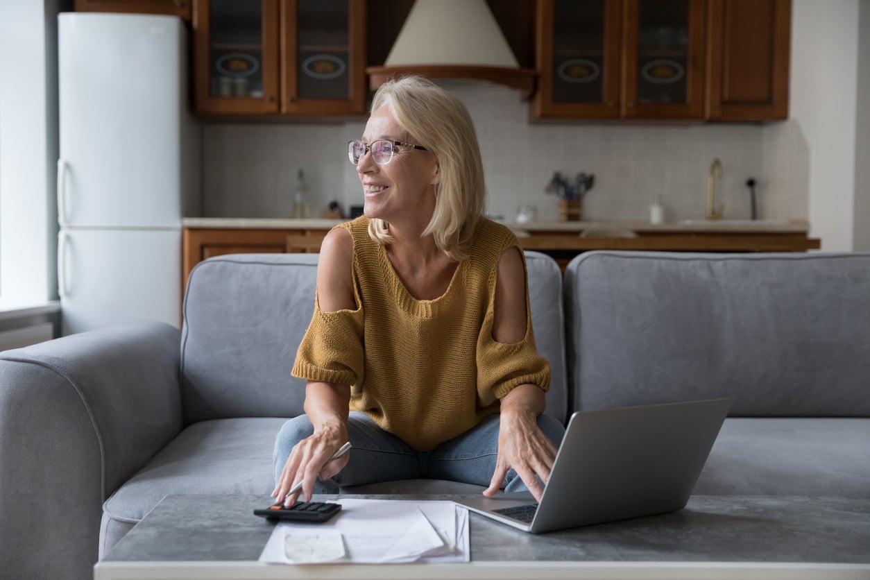 Smiling woman with glasses sits on a couch using a laptop and calculator, with papers on the table in front of her, in a cozy kitchen setting—a reflection of how Women Are Changing Their Spending Behaviors.