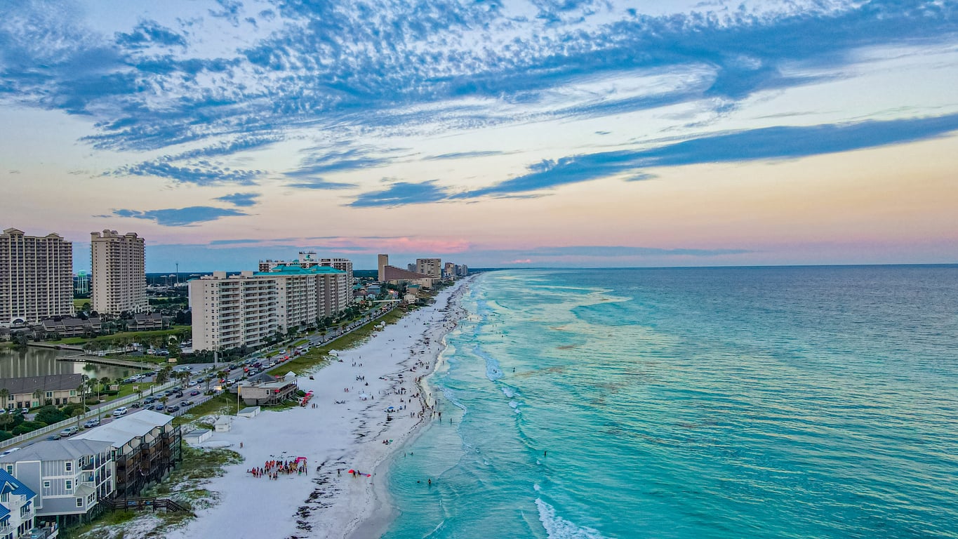 Aerial view of a beach with turquoise water, people on the white sand, and high-rise buildings along the shoreline under a blue and pink evening sky—one of the most stunning travel destinations for those seeking unforgettable coastal beauty.