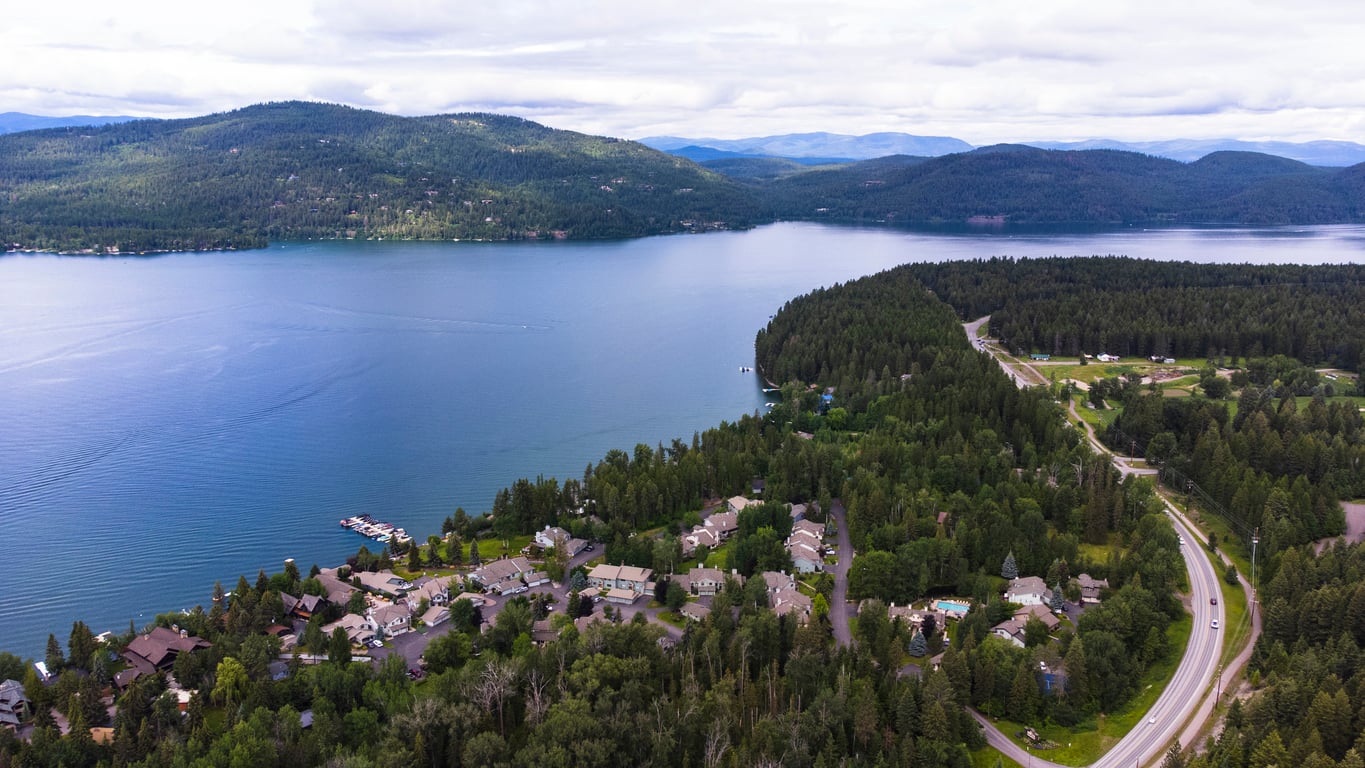 Aerial view of a lakeside community surrounded by dense forests and hills, with a winding road and docks along the shoreline under a cloudy sky.