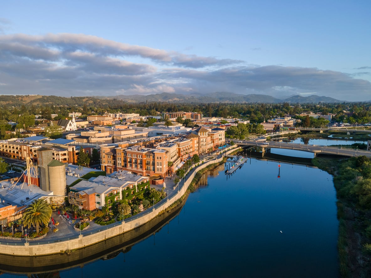 Aerial view of a small city by a calm river, with buildings, a waterfront walkway, a bridge, green trees, and hills under a partly cloudy blue sky.