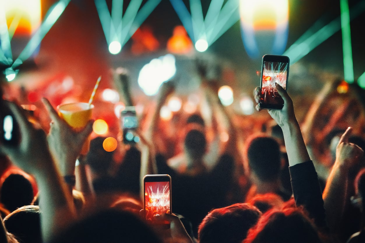 A crowd at a concert holds up phones to record the stage, with colorful lights and lasers shining in the background—a stark contrast to life before smartphones, when hands were raised in excitement without screens, and memories lived only in the moment. Some people also hold drinks as the energy surges all around.