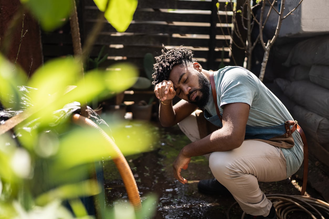A man squats outdoors, wiping sweat from his forehead, looking tired. He wears a blue apron and light pants, surrounded by gardening tools and plants—a quiet moment that reflects Baby Boomer beliefs in hard work and dedication. Sunlight filters through leaves nearby.