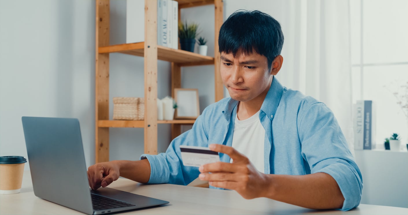 A man sitting at a desk looks worried while holding a credit card and using a laptop, possibly concerned about an online purchase. Bookshelves and a coffee cup are visible in the background.