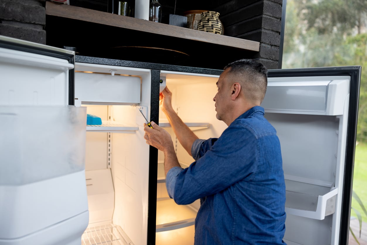 A man in a blue shirt uses a screwdriver to repair or adjust the door of an open refrigerator in a kitchen, inspecting the mostly empty shelves for any missing products. Both the refrigerator and freezer doors are open.