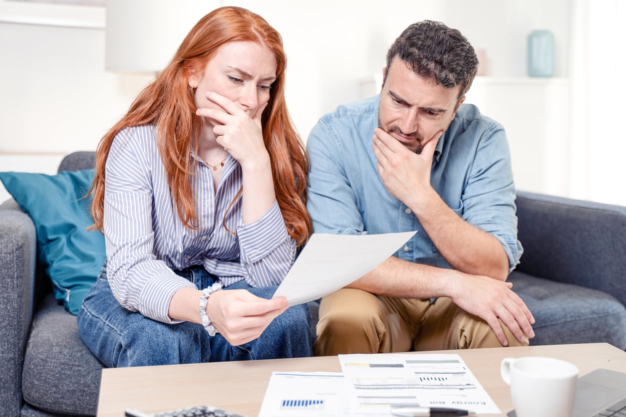A worried couple sits on a couch, examining a piece of paper with charts and documents spread on the table in front of them, suggesting financial concerns.