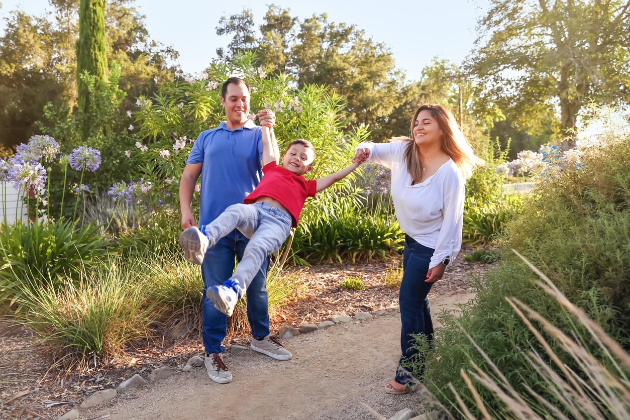 A smiling child is swung playfully by two adults, one on each side, as they walk together on a garden path surrounded by greenery and flowers on a sunny day—a moment that captures the joy in experiences and memberships worth paying for.