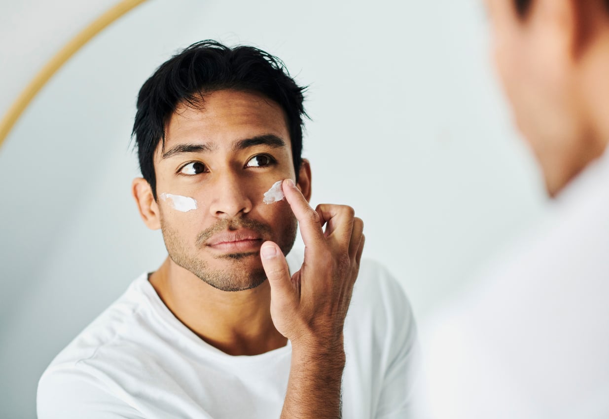 A man in a white shirt, practicing money saving habits, applies moisturizer to his face while looking at himself in a mirror.