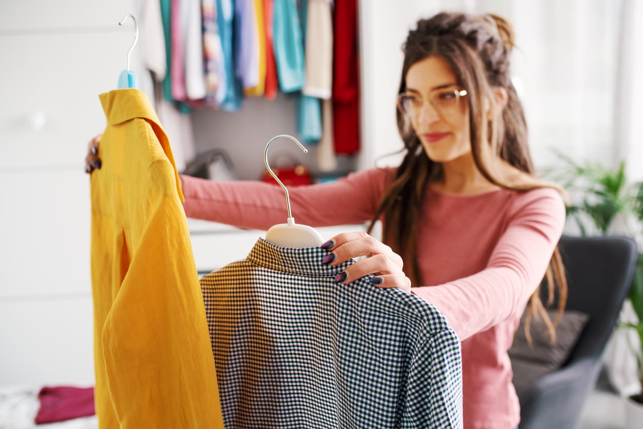A woman stands in a room holding up a yellow shirt and a checkered shirt on hangers, appearing to decide between them—a small moment that reflects how Women Are Changing Their Spending Behaviors. Clothing is visible in the background.