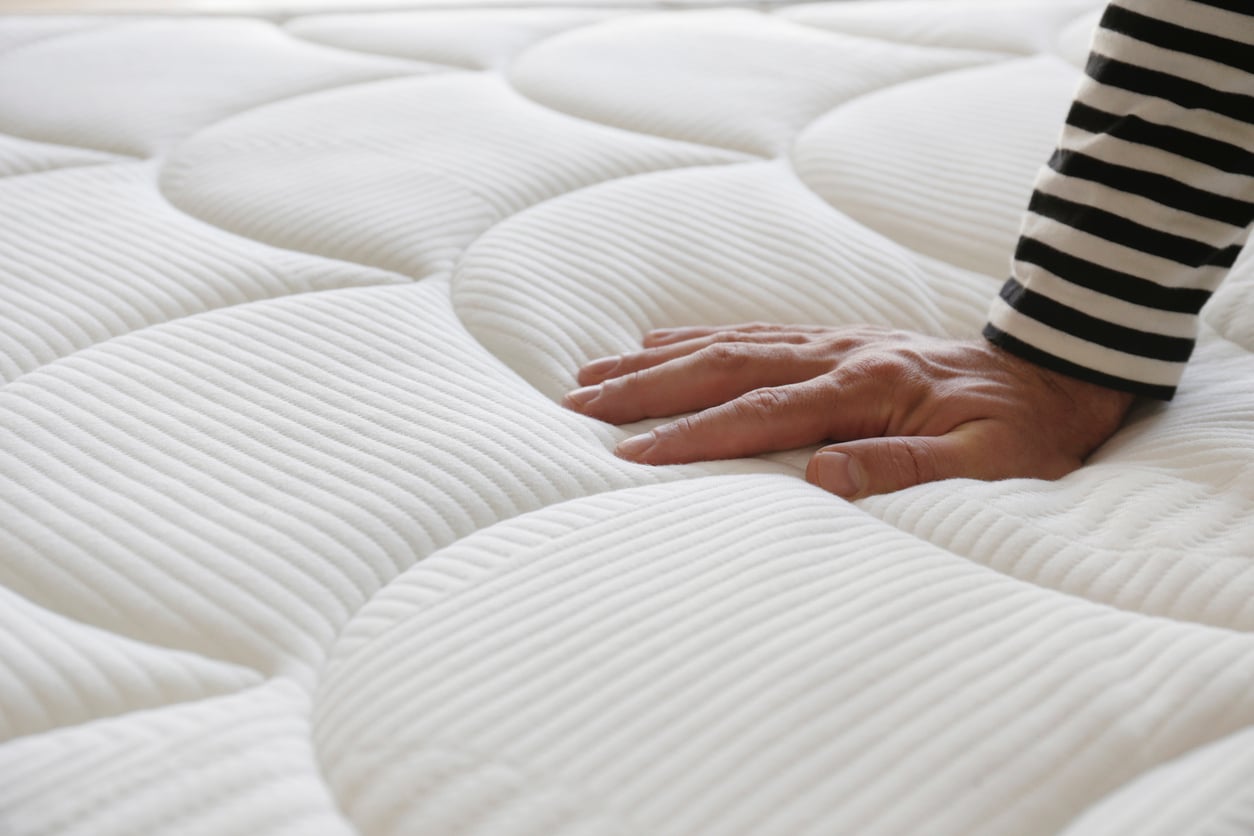 A hand presses down on a white, quilted mattress, testing its softness—unlike products that don't last, this quality piece promises enduring comfort. The person wears a long-sleeve shirt with black and white stripes.