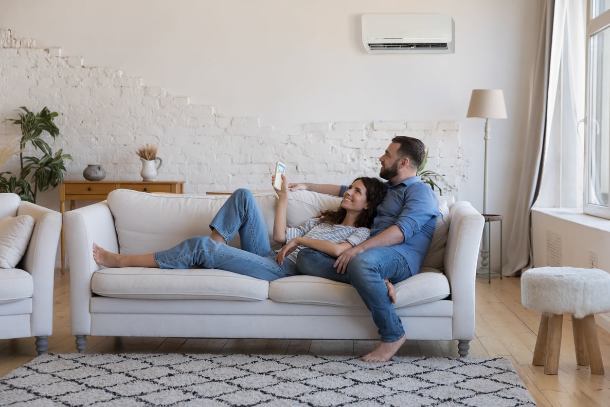 A couple relaxes on a white sofa in a bright living room, smiling and looking at a smartphone together. The room has plants, a rug, large windows, and an air conditioner on the wall.