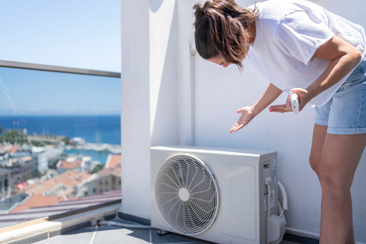 A woman in a white t-shirt and denim shorts gestures towards an outdoor air conditioning unit on a balcony, with a scenic coastal town and ocean visible in the background.